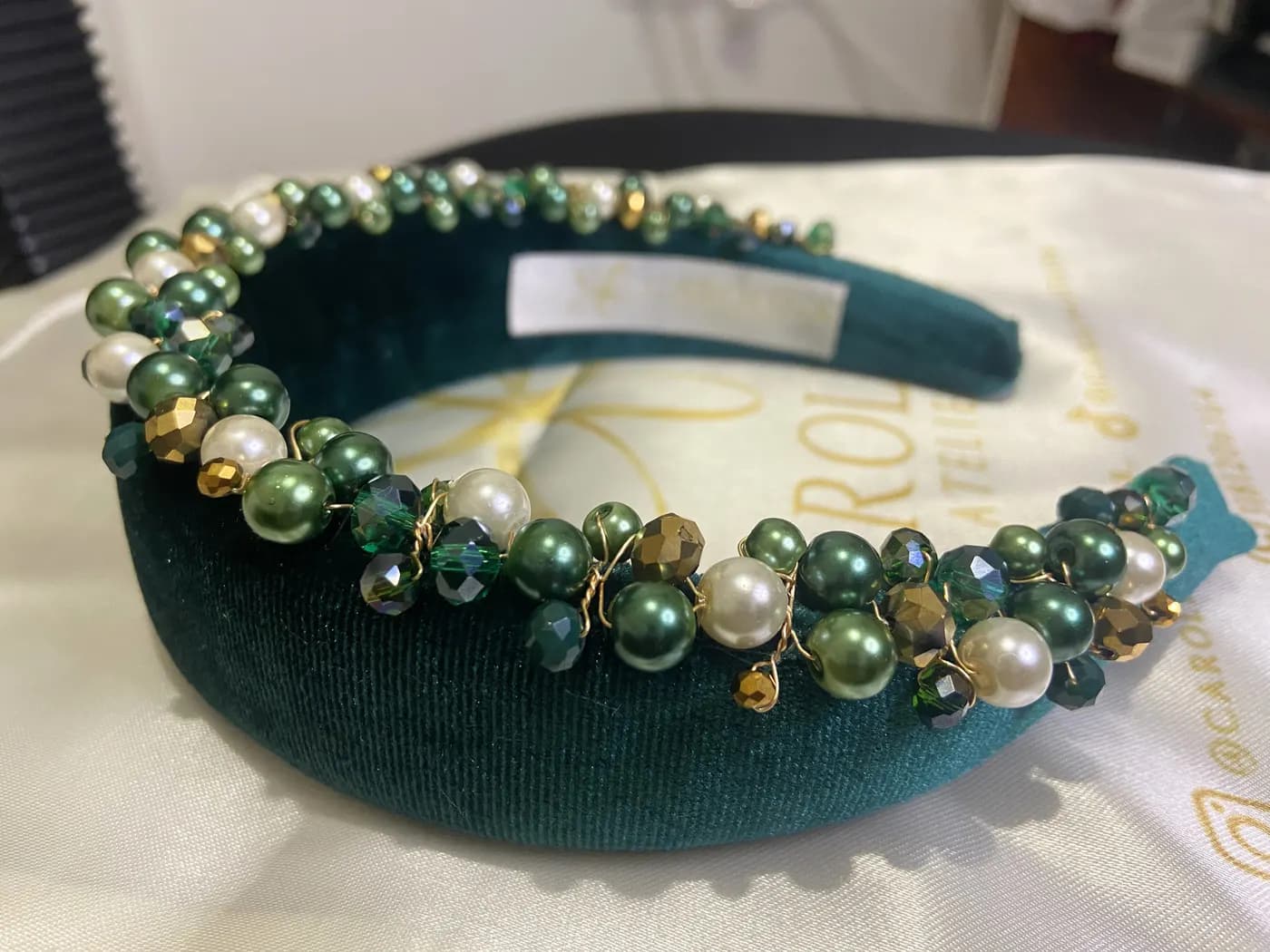 Diadema Loft de terciopelo verde esmeralda con perlas y cristales verdes y dorados tejidos a mano, sobre su bolsa de satín